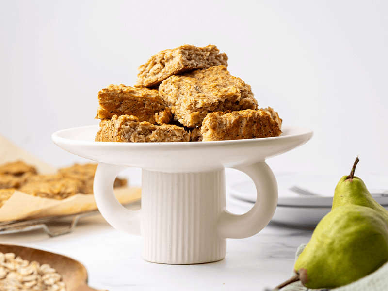 Plate of homemade fruit and oat bars stacked on a white cake stand, with fresh pears and rolled oats in the background, perfect for a healthy snack or lunchbox treat.