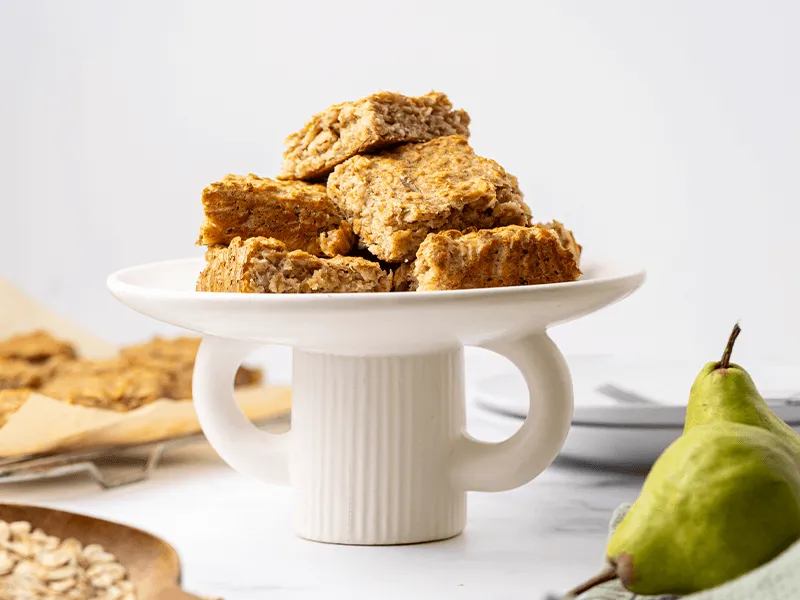 Plate of homemade fruit and oat bars stacked on a white cake stand, with fresh pears and rolled oats in the background, perfect for a healthy snack or lunchbox treat.