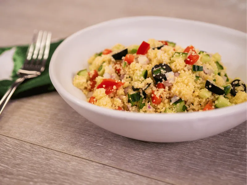 Greek-style couscous salad with cucumber, tomato, red onion, and black olives tossed in a lemon‑olive oil dressing, served in a white bowl.
