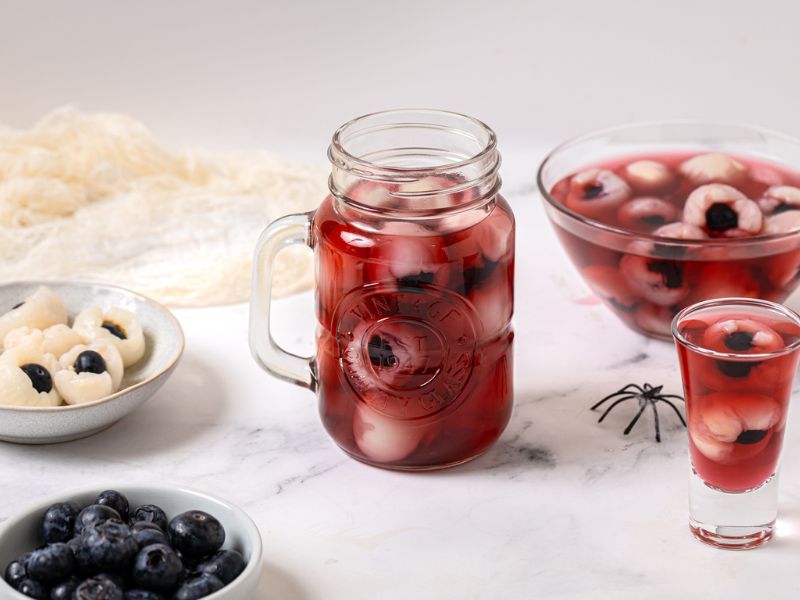 Halloween lychee eyeballs drink with blueberries in cranberry juice, served in a mason jar and glass, surrounded by fresh lychees and blueberries on a white marble background