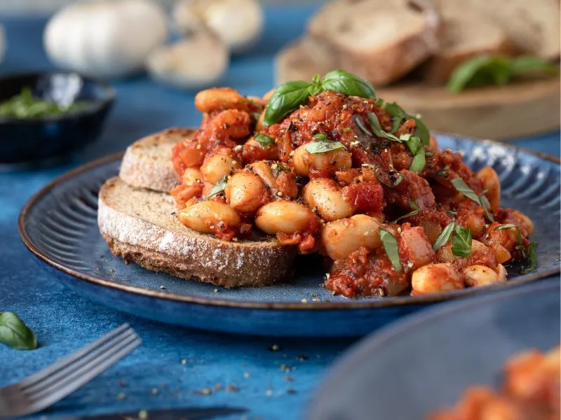 A white plate on a wooden table contains two slices of sourdough bread, toasted, and topped with a rich, cooked tomatoes and bean mixture. The whole meal has been liberally sprinkled with chopped parsley.