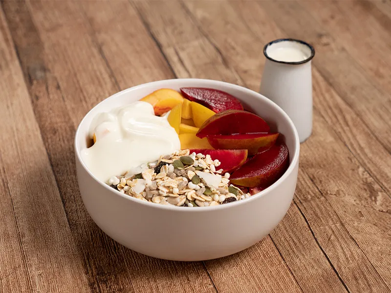 A white bowl sits on a wooden table top with small jug of milk alongside it. Inside the bowl is natural muesli, slices of plum and peach with a large dollop of natural yoghurt to one side.