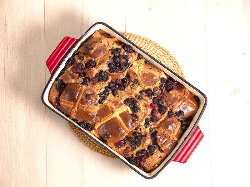 A top-down view from above shows a red baking dish on a wooden table top. Inside the baking dish is a bread and butter pudding made with hot cross buns, clearly visible, and a scattering of purple berries.
