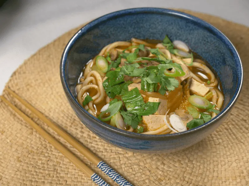 Kimchi, tofu, bok choy, and noodle soup in a blue bowl—spicy kimchi broth with udon noodles, tofu cubes, bok choy, and scallions, garnished with cilantro.