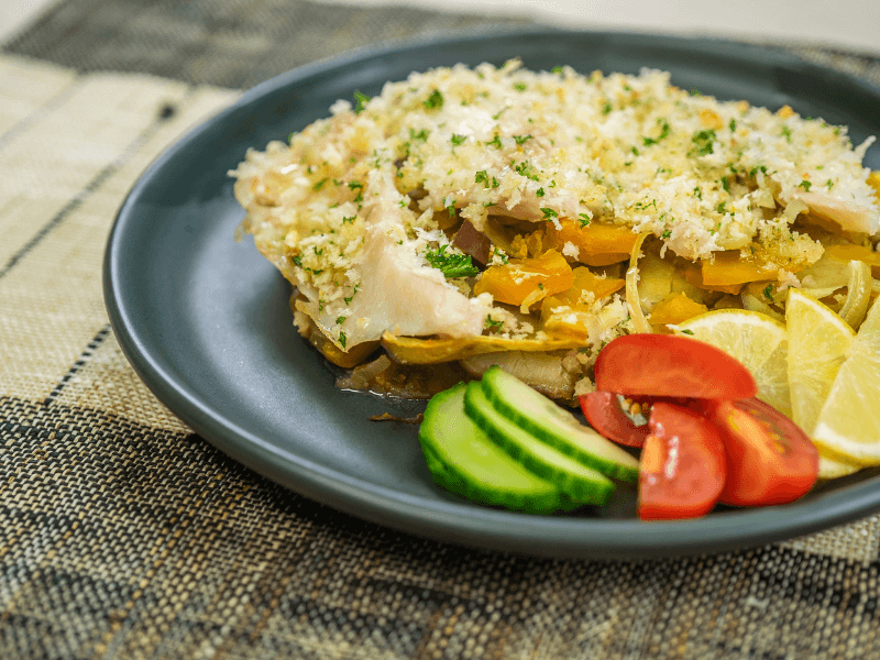 Lemon‑crusted baked fish served over a kumara and pumpkin bake, topped with herbed breadcrumbs, with cucumber, cherry tomatoes, and lemon wedges on the side.