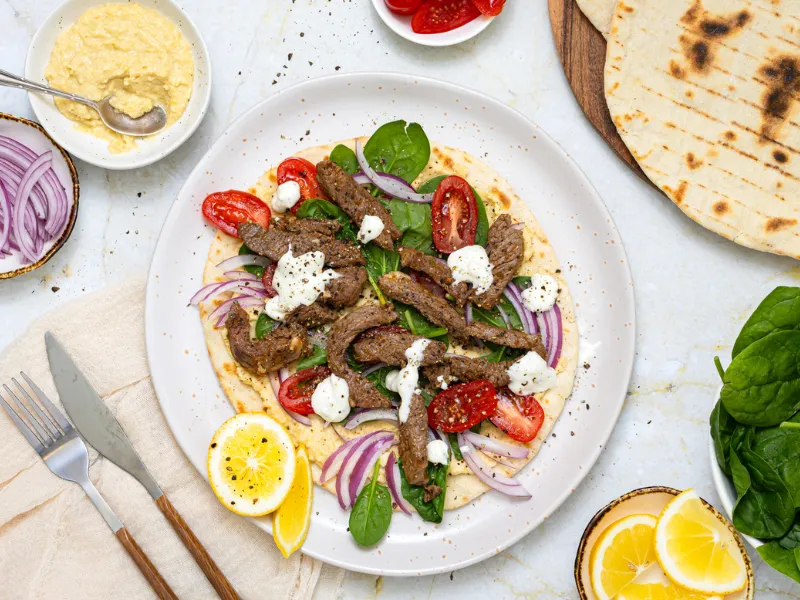 Overhead view of Greek lamb souvlaki wrap on flatbread with spinach, tomato, red onion and yoghurt sauce, served with hummus, lemon wedges and extra flatbreads