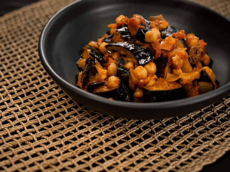 Lebanese chickpea and eggplant stew (maghmour) in a tomato‑garlic sauce, with tender aubergine pieces and onions in a black bowl.