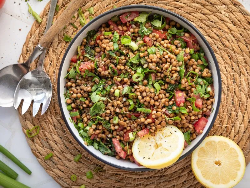 Bowl of lentil salad with chopped tomatoes, fresh herbs, and sliced green onions, garnished with black pepper and served with lemon halves on a woven placemat, with salad servers on the side.