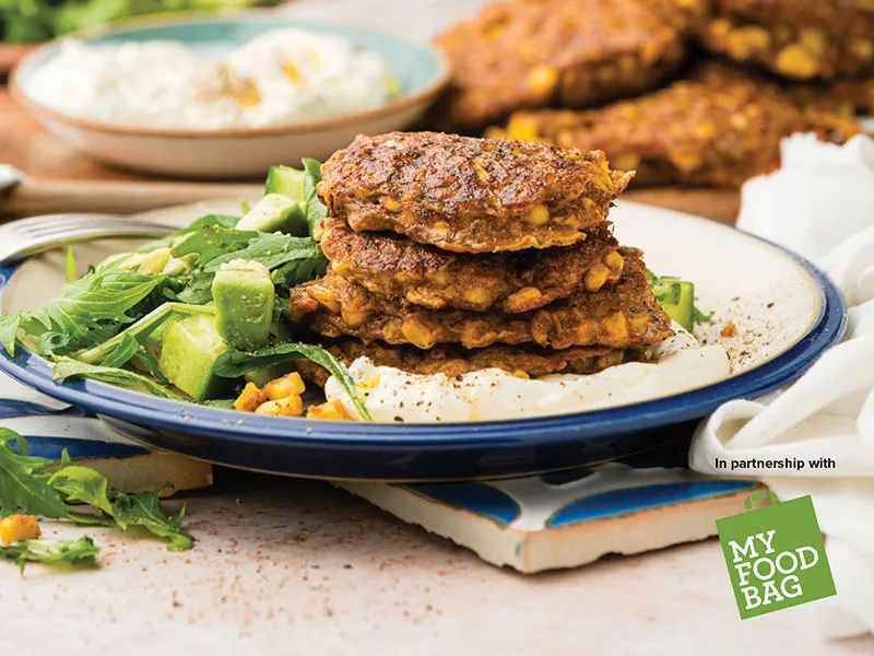 Image shows a plate stacked with 3 large round corn fritters, accompanied by a fresh cucumber salad and a dollop of feta whip.