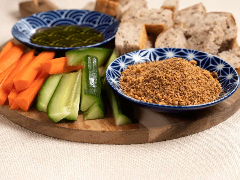 Moroccan dukkah—crunchy nut and spice blend featuring toasted nuts (like almonds or hazelnuts), sesame, cumin, coriander, and pepper—served as a dip with olive oil, bread, and fresh veggies.