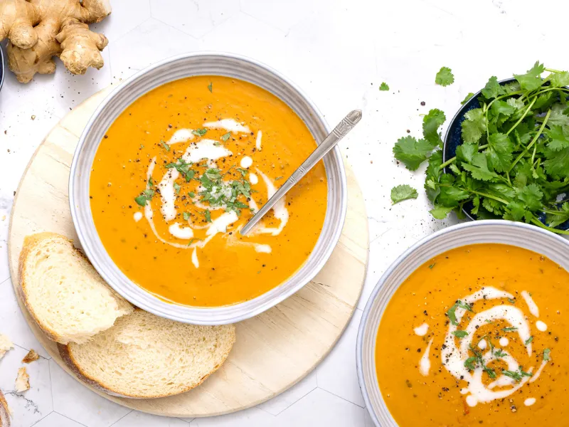 Bowl of creamy orange mulligatawny lentil soup garnished with yogurt swirl and fresh coriander, served with sliced bread on a wooden board