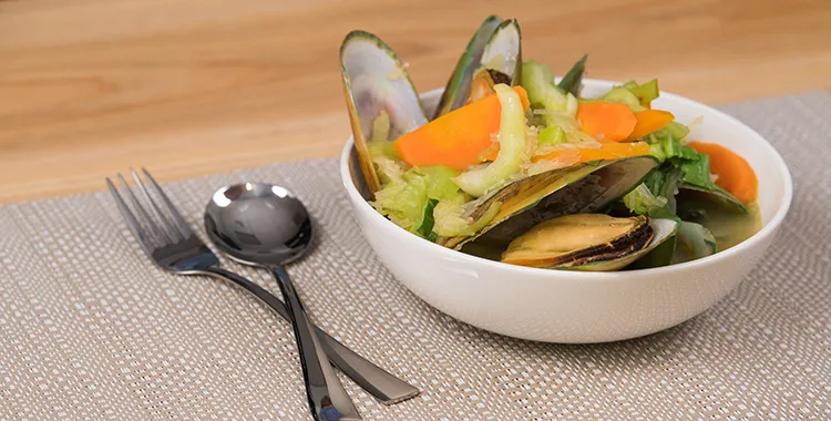 Steamed mussels with bok choy and carrot slices in a light broth, served in a white bowl on a neutral placemat with fork and spoon.