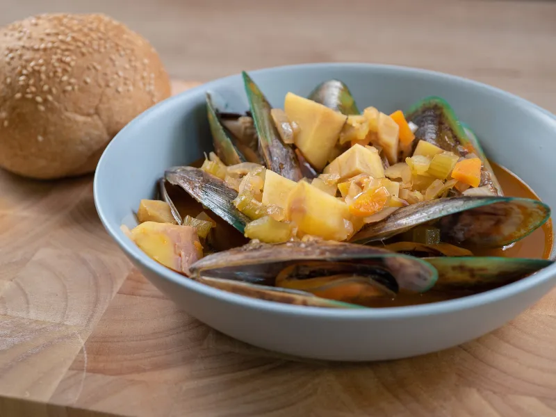 Bowl of mussel and kūmara stew with carrot and celery, served with a wholemeal roll.