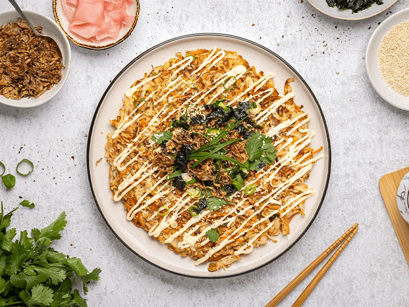 Crispy Japanese okonomiyaki cabbage pancake topped with creamy mayonnaise, fresh green onions, seaweed, sesame seeds, and garnished with fried shallots and cilantro, surrounded by pickled ginger, extra toppings, and chopsticks on a marble table setting.