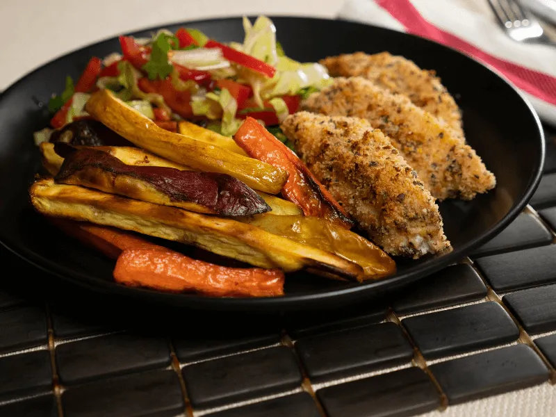 Oven-baked fish and chips: crumbed baked fish fillets with kumara (sweet potato) and carrot chips, served alongside a fresh salad of lettuce, capsicum, and herbs on a black plate.