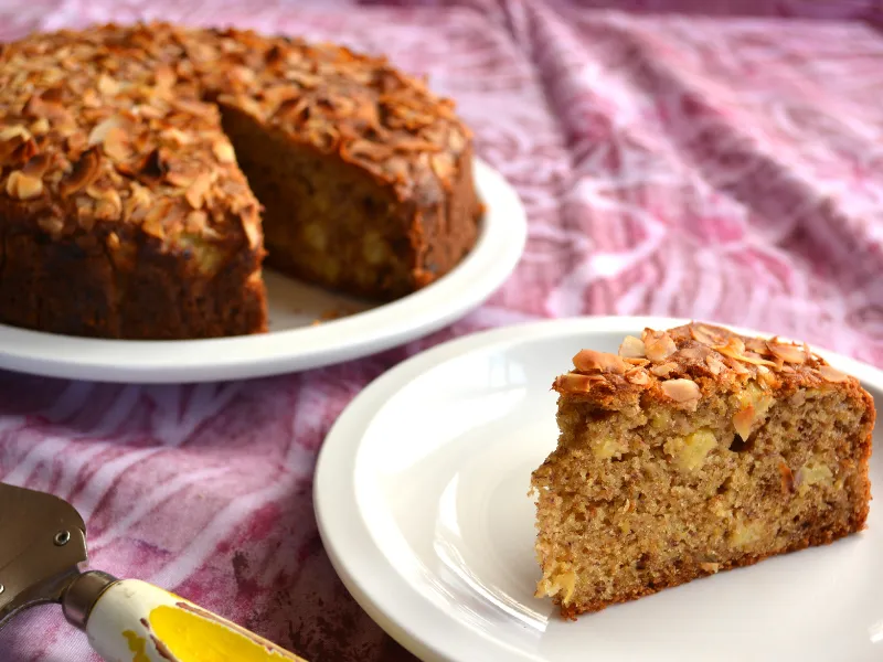 Moist Pacific fruit cake topped with toasted almonds on a white plate, slice served in front, tropical dessert recipe for island-style baking and celebrations.