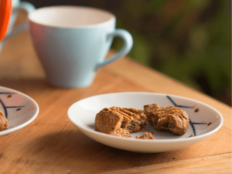 Peanut butter and banana cookies with fork marks, one broken on a small white plate beside pastel mugs on a wooden table.