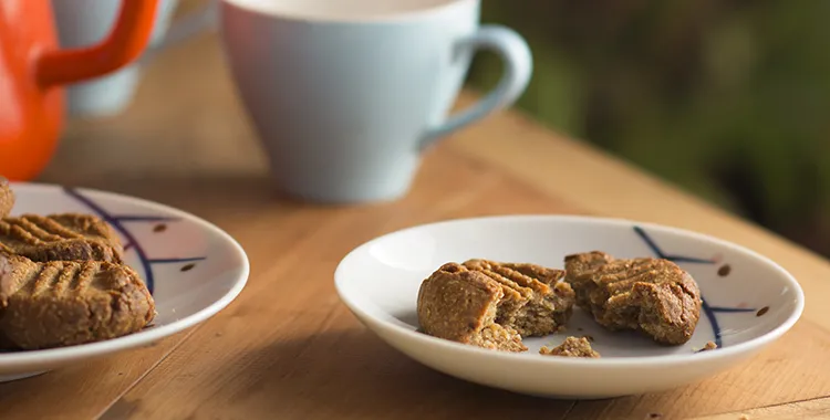 Peanut butter and banana cookies with fork marks, one broken on a small white plate beside pastel mugs on a wooden table.