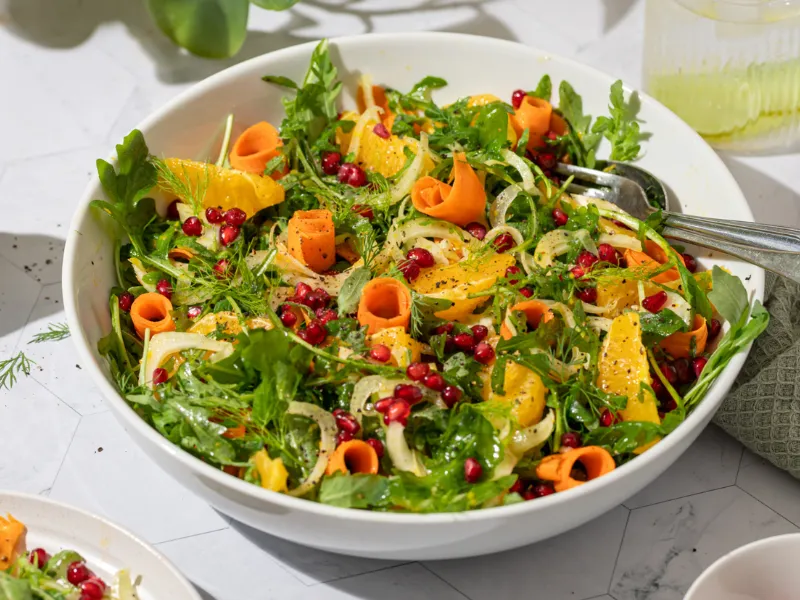 A large white bowl filled with a vibrant salad of thinly sliced fennel, carrot ribbons, baby rocket, orange segments and scattered pomegranate seeds, garnished with fennel fronds and served with a light citrus dressing, styled on a bright tabletop.