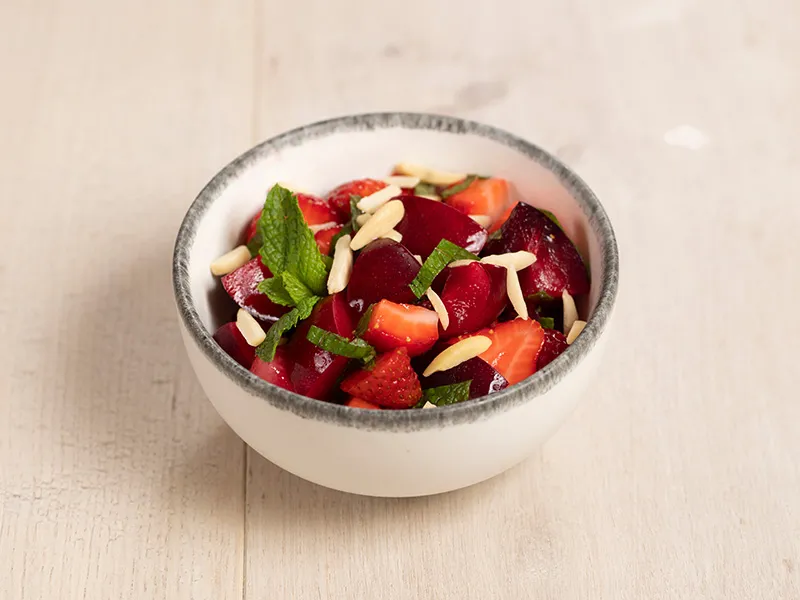 A white bowl with grey edging on an off-white surface holds a serving of plum, strawberry and almond salad