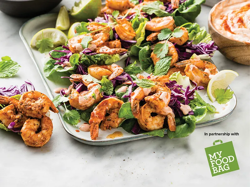 Quick, easy, and so delicious, these prawn tacos by My Food Bag are sure to be a big hit this summer. This image shows grilled prawns spilling out from a lettuce cup with shredded red cabbage, lime quarters and coriander leaves.