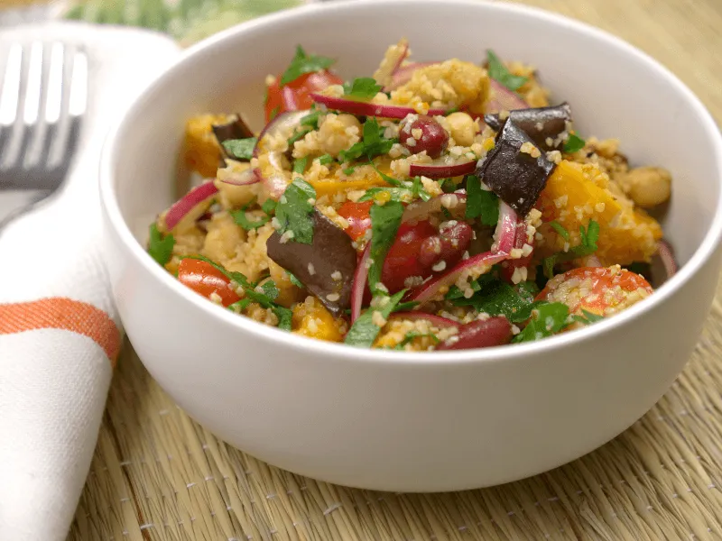 Roasted pumpkin, eggplant, and bean salad with couscous, red onion, cherry tomatoes, and parsley in a lemony dressing, served in a white bowl.