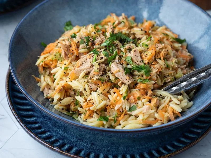 Bowl of orzo pasta salad with shredded carrot, flaked tuna, chopped herbs, and cracked black pepper, served in a blue ceramic dish with a fork.