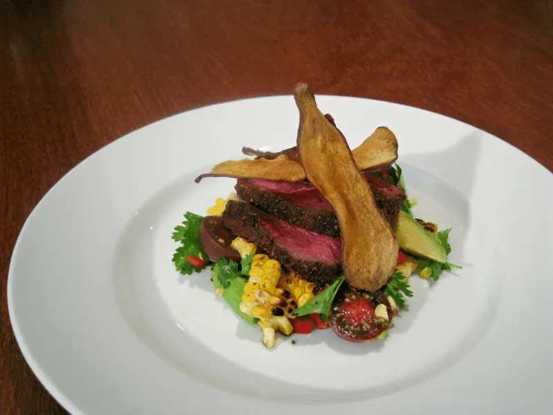 Sliced roast venison on a Mexican-style corn and tomato salad with avocado and cilantro, garnished with crisp vegetable chips on a white plate.