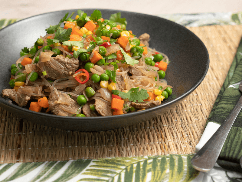 Sapasui (Samoan chop suey) with beef, glass noodles, peas, carrot, corn, green beans, and spring onion dressed in soy sauce and garnished with fresh coriander in a black bowl.