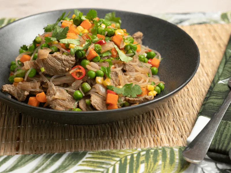 Sapasui (Samoan chop suey) with beef, glass noodles, peas, carrot, corn, green beans, and spring onion dressed in soy sauce and garnished with fresh coriander in a black bowl.
