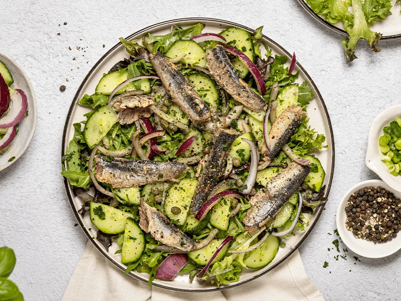 Summer sardine salad with mixed leafy greens, sliced cucumber, red onion, capers, and herbs on a white plate, overhead view of healthy Mediterranean-style seafood salad on a light background.