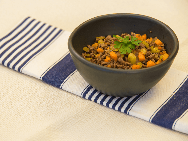 Savoury mince with diced carrots and celery in a black bowl, garnished with parsley, served on a blue‑striped napkin.