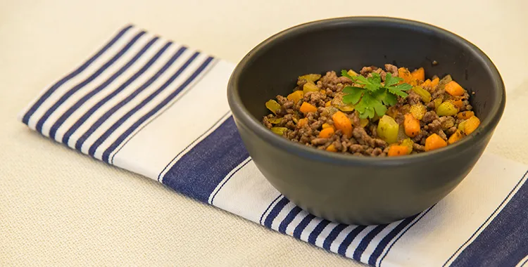 Savoury mince with diced carrots and celery in a black bowl, garnished with parsley, served on a blue‑striped napkin.