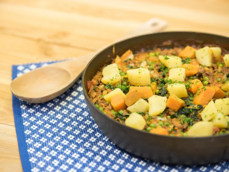 Pan of savoury vegetables and mince topped with diced kūmara and potatoes on a blue mat.