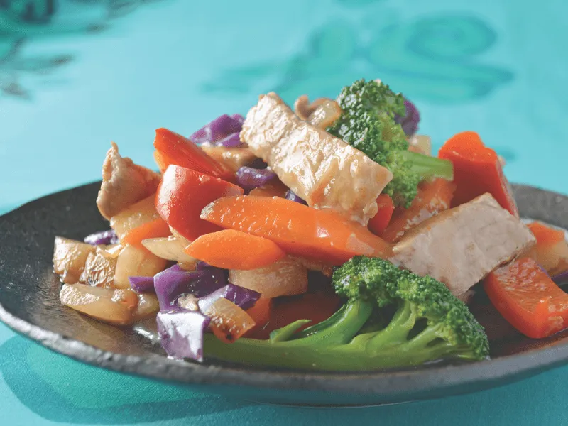 Simple sweet and sour pork with strips of lean pork, carrots, red capsicum, broccoli, onion, and purple cabbage in a tangy sweet and sour sauce, served on a dark plate.