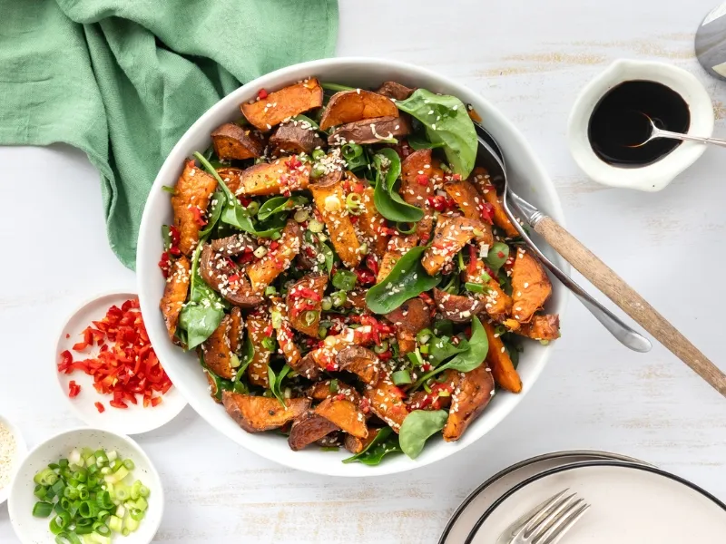 Roasted kumara (sweet potato) salad with fresh spinach leaves, chopped red chili, spring onions, and sesame seeds, served in a white bowl with a dressing on the side.