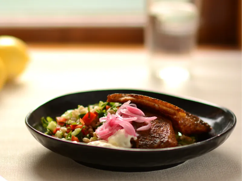 Spiced fish fillets served with barley salad, yogurt and pink pickled onions on a black plate.