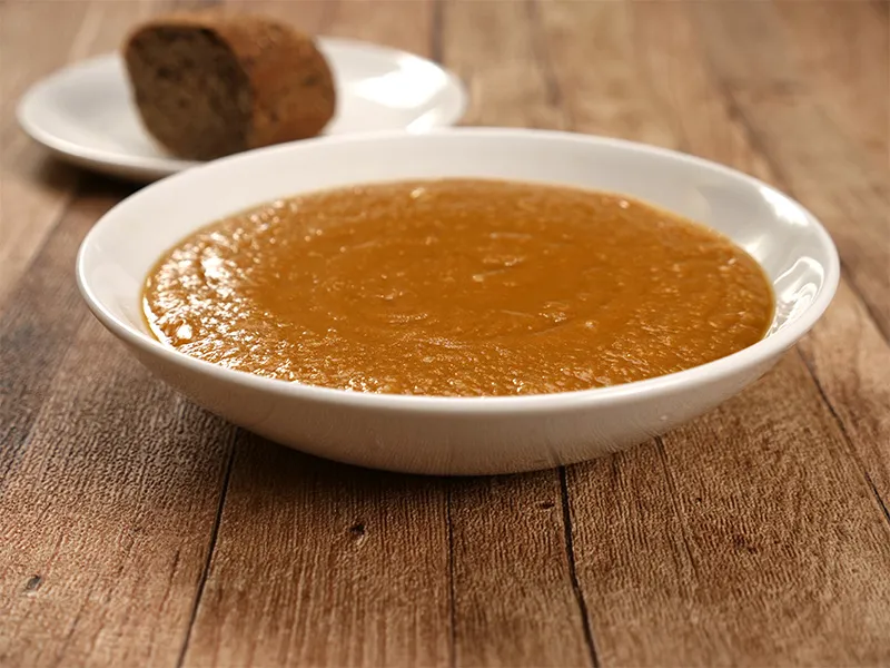 A shallow white bowl on a brown, wood surface holds a serving of spicy red lentil soup with a portion of brown bread in the background on a white plate.