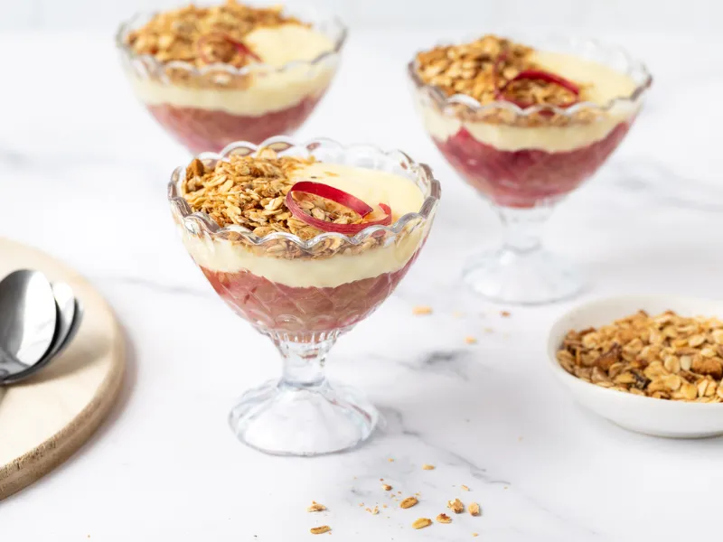 Rhubarb and custard parfaits topped with crunchy granola, served in elegant glass dessert dishes on a white marble surface.