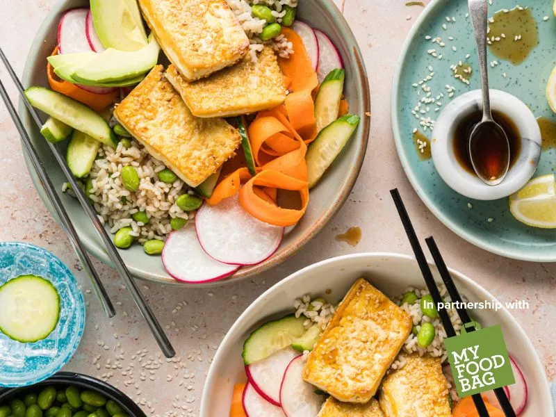 Overhead view of colourful summer tofu poke bowls with brown rice, edamame, avocado, cucumber, carrot ribbons and radish, drizzled with lime ponzu dressing, styled on a light table with chopsticks and sesame seeds in partnership with My Food Bag.