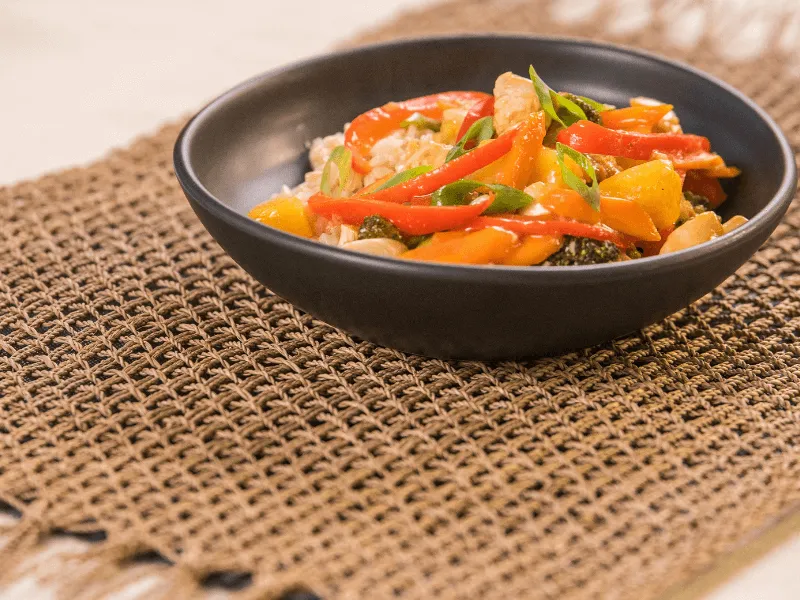 Sweet and sour chicken stir‑fry with red and yellow bell peppers and pineapple over rice, garnished with sliced spring onions in a black bowl.