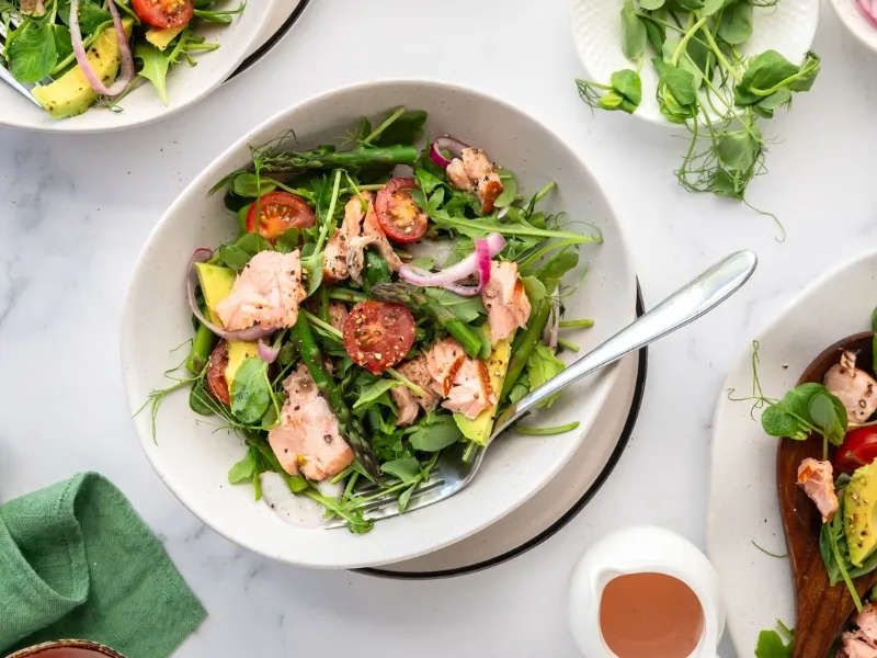 Bowl of fresh salmon salad with arugula, cherry tomatoes, avocado, red onion, and asparagus, served with a fork on a white marble surface.