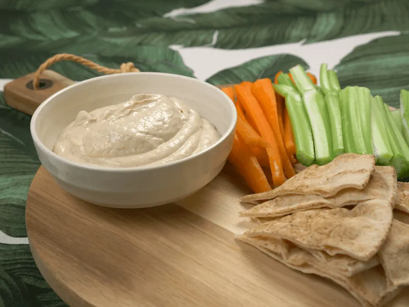 Tofu and tahini dip served with pita wedges, carrot sticks, and celery on a wooden board.