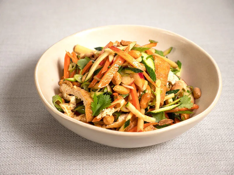 A white bowl filled with tofu salad sits on a beige background. Inside the bowl, we can see strips of fried tofu, carrot and courgette juliennes, apple slices, spring onions and peanuts with a sprinkling of chopped coriander on top.