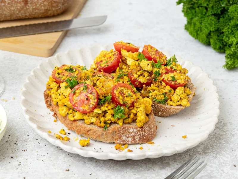 Scrambled tofu toast with cherry tomatoes and parsley on whole grain bread served on a white plate.