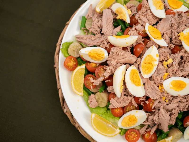 Tuna, egg, and green banana salad with cherry tomatoes, lettuce, and green beans, garnished with lemon wedges on a platter.