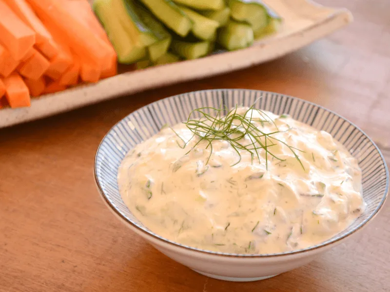 Creamy tzatziki yogurt dip with cucumber and dill in a bowl, garnished with fresh dill, with sliced carrots and cucumbers in the background.