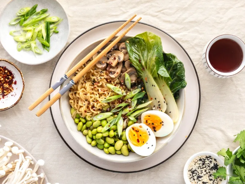 Bowl of ramen noodles topped with halved soft-boiled egg, edamame, bok choy, sliced mushrooms, and green onions, garnished with sesame seeds and chili flakes, served with chopsticks.