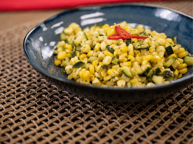 Warm courgette and corn salad with diced zucchini, sweet corn kernels, fresh herbs, and a garnish of sliced red chili in a blue bowl.