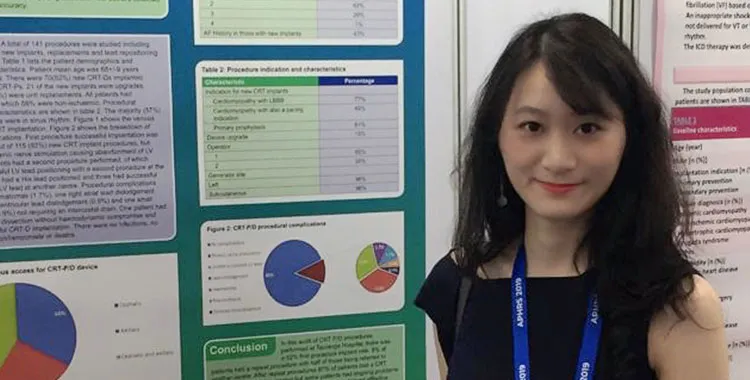 Young, Chinese woman Becky Liao, standing in front of a large colourful poster displaying graphs and statistical medical information